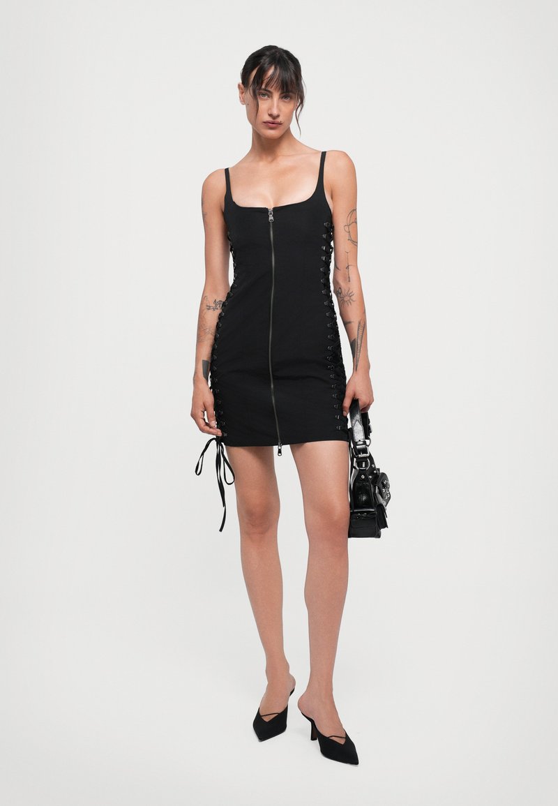 Svart ärmlös klänning med dragkedja fram och snörningsdetaljer på sidorna. Tyget är slätt och designen är figurnära.