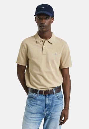 Mand iført en beige kortærmet polo shirt, blå jeans med brunt bælte og en marineblå baseball cap, står op af en ensfarvet baggrund.