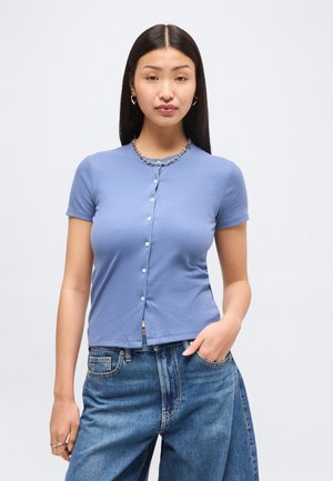 Žena s dlhými čiernymi vlasmi, oblečená v svetlomodrej krátkym rukávom zapínanej košeli a voľných modrých džínsoch, stojaca s jednou rukou v vrecku.