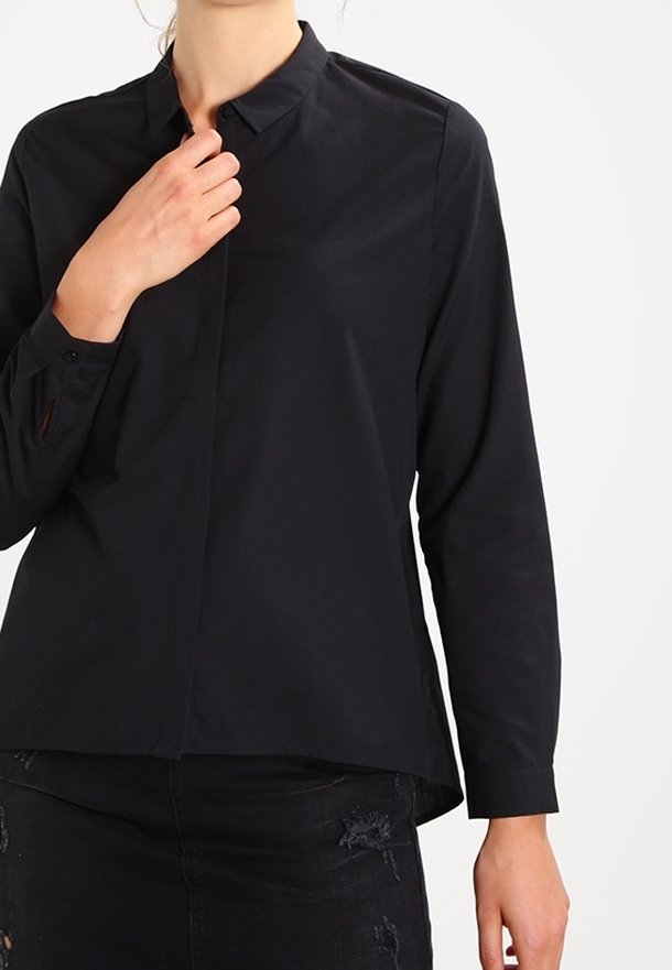Femme portant une chemise noire à manches longues avec col et une jupe noire usée, avec une main touchant le col.