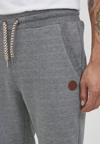 Grå strikkede joggebukser med en teksturert finish, som har et gummimerke og brune snoraksenter. Sidelommene er synlige.
