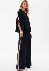 Zwarte broek met wijde pijpen en een beige zijkantstreep, gecombineerd met een zwarte, losvallende top. Soepele stof, ontspannen ontwerp en minimalistische accenten.