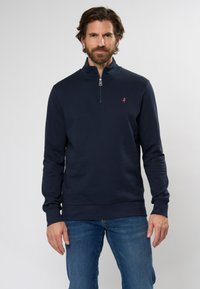 Sudadera con cremallera de color azul marino hecha de tejido suave, que cuenta con un alto cuello, puños acanalados y un pequeño logotipo en contraste en el pecho.