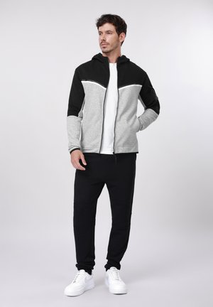 Mees, kes kannab musta ja halli lukuga dressipluusi, valget t-särki, musti pükse ja valgeid tenniseid, seisab ühe käega taskus ühtlase tausta ees.