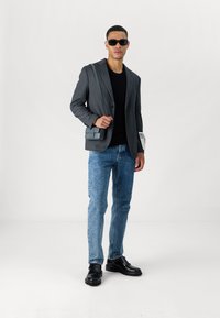 Pilkas pritaikytas švarkas, juoda marškiniai, šviesiai mėlynos džinsai, juodi batai ir pilkas krepšys per petį. Modelis dėvi juodus akinius nuo saulės. Lygi tekstūra.