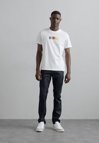 T-shirt en coton blanc avec un logo "SANDRO" coloré sur la poitrine, associé à un jean en denim bleu foncé et des baskets blanches. Design simple et moderne.