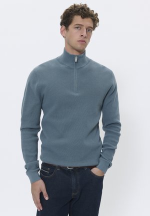 Dolga rokavica, svetlo modra zip-up jopica z teksturirano tkanino in visokim ovratnikom. Skupaj z temnimi jeans hlačami in rjavim pasom.