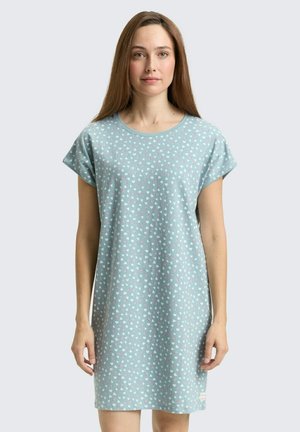 Vrouw draagt een lichtblauwe jurk met korte mouwen met kleine witte en roze hartjespatronen, staand tegen een effen lichte achtergrond.