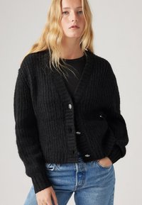 Μαύρο ριγέ κάρντιγκαν με V-λαιμόκοψη, υφασμάτινη υφή και διακοσμητικά κουμπιά. Φοριέται πάνω από μαύρη μπλούζα και συνδυάζεται με μπλε τζιν.