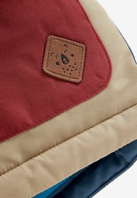 Gros plan sur un tissu avec des sections rouges, beiges et bleues et un patch marron cousu présentant un logo en forme de tête d’ours.