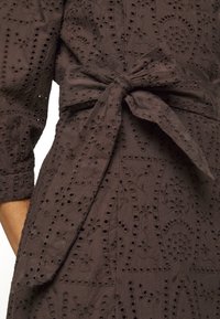 Robe en tissu œillet marron avec une broderie florale complexe et une ceinture nouée à la taille, montrant une partie de la manche et du torse.