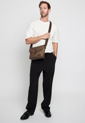 Homme portant une chemise blanche à manches courtes, un pantalon noir, des chaussures noires, tenant un sac porté en bandoulière marron avec une sangle sur l'épaule, debout sur un fond gris.