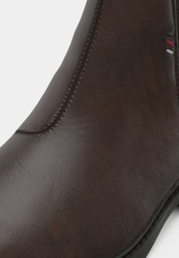 Primo piano di un boot in pelle marrone con cuciture visibili e un piccolo dettaglio ricamato di tre strisce orizzontali in blu navy, rosso e bianco.