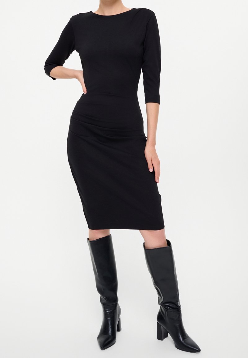 Femme portant une robe noire ajustée au genou avec des manches trois-quarts et des bottes noires à talons montant jusqu'aux genoux, sur un fond uni.