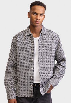 Man met kort krullend haar die een gemusterde blouse over een witte tanktop en zwarte broek draagt, met een hand in de zak, op een neutrale achtergrond.