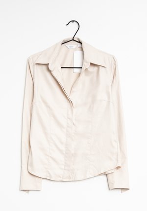 Camisa de manga larga beige con cuello, que presenta una textura suave, un corte entallado y un patrón de rayas verticales. Cuelga de un gancho negro.