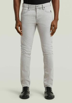 Mann trägt schlank geschnittene hellgraue Jeans und schwarze Lederschuhe und steht vor einem schlichten hellen Hintergrund.