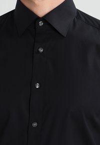 Black button-up shirt made of smooth fabric with a classic collar. Features silver buttons and a streamlined fit, lacking patterns or embellishments.