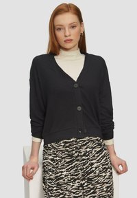 Schwarze Strickjacke mit V-Ausschnitt, ausgestattet mit vier Knöpfen und geripptem Muster, getragen über einem cremefarbenen Top und einem gemusterten Rock.