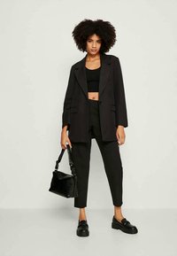 Sieviete ar cirtainiem matiem, tērpusies melnā žaketē, melnā kroptopā, melnās biksēs un melnās kurpēs, tur melnu somiņu pret viendabīgu fona.