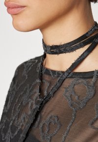 Gros plan sur le cou d'une personne portant un top noir transparent avec des motifs floraux texturés et un collier ras-du-cou noir effiloché noué autour du cou.