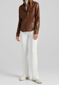 Giacca camicia in finta pelle marrone con due tasche sul petto, chiusura con bottoni e colletto, abbinata a pantaloni bianchi a coste e scarpe beige.