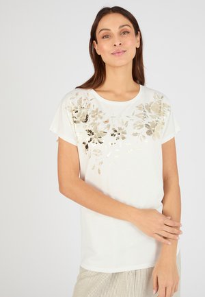 Frau mit langen braunen Haaren, die ein weißes kurzärmeliges Oberteil mit goldenen floralen Pailletten und hell gestreifte Hose trägt, steht vor einem einfarbigen Hintergrund.