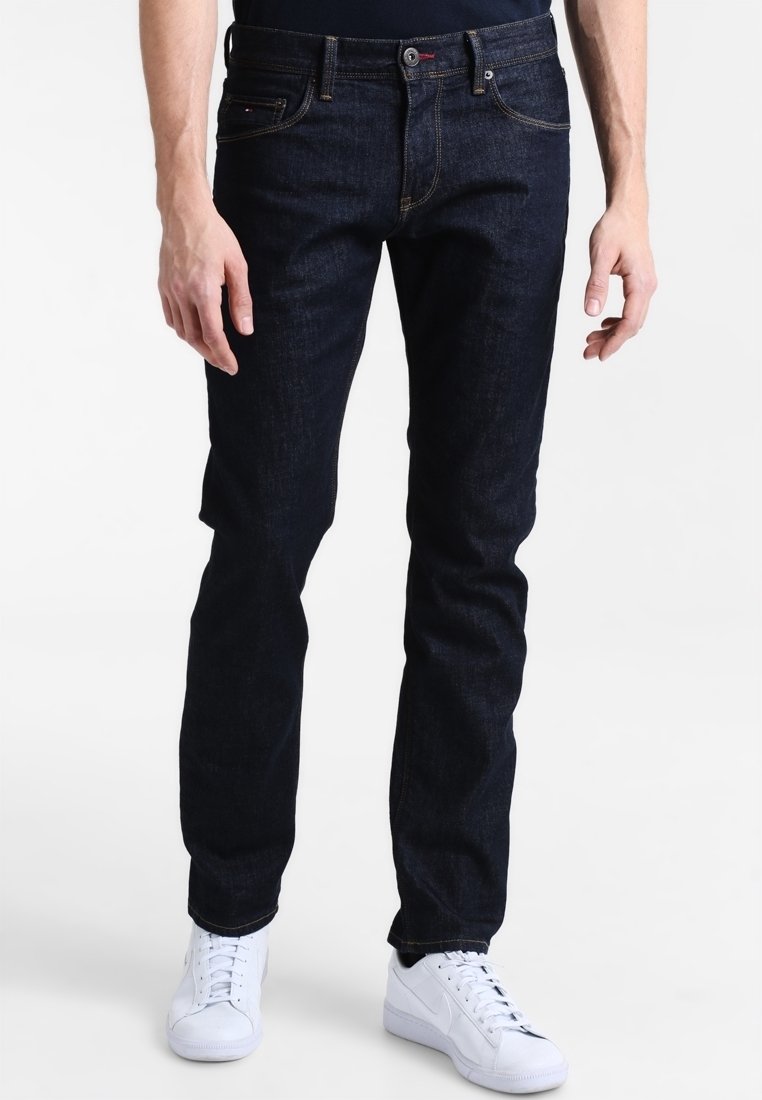 Navy blue denim jeans with a slim fit, contrasting stitching, five-pocket design, finished with a subtle logo and paired with white sneakers.