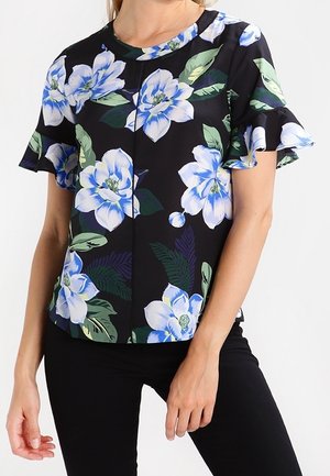 Vrouw draagt een zwarte blouse met korte mouwen met een grote blauw-witte bloemenprint en ruches aan de mouwen, gecombineerd met een zwarte broek.