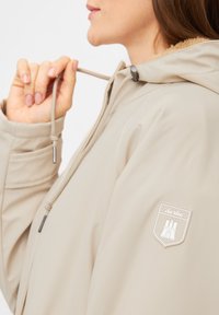 Beige weather-resistant jacket with a drawstring hood, featuring a soft fur lining and a logo patch on the sleeve.