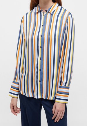 Kobieta nosząca koszulę z długim rękawem zapinaną na guziki z pionowymi paskami w kolorach niebieskim, białym, pomarańczowym i żółtym, zestawioną z ciemnoniebieskimi spodniami.