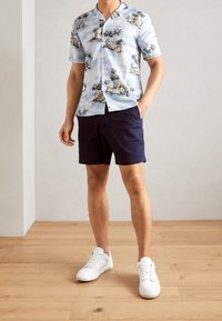 Chemise légère à manches courtes de couleur bleu clair avec un imprimé de palmier et de planche de surf, assortie à un short navy et des baskets blanches sur un sol en bois.