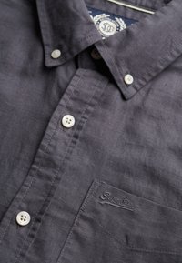 Camisa de botones gris oscuro con cuello, botones blancos, un bolsillo en el pecho con un logo bordado y un acabado de tela texturizada.