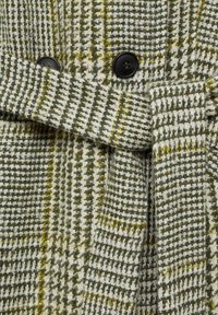 Manteau en laine à motif pied-de-poule vert et blanc avec des boutons noirs et une ceinture assortie en tissu nouée à la taille.