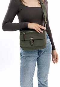 Olivově zelená crossbody kabelka s kapsou na zip, klopou uzavíratelnou hnědým sponou a nastavitelným popruhem, vyrobená z odolné tkaniny.