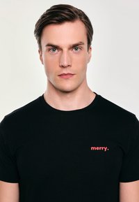 Černé bavlněné tričko s krátkým rukávem, které má červený textový grafický prvek s nápisem "merry" umístěný na levé straně hrudi.