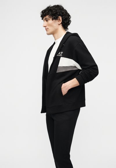 Νεαρός με κατσαρά μαλλιά που φοράει μαύρο φούτερ με κουκούλα, με λευκές και γκρι ρίγες, και μαύρο παντελόνι, όρθιος σε προφίλ με τα χέρια στις τσέπες.