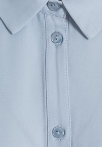 Col de chemise bleu clair avec trois boutons bleus assortis cousus le long du pied de la patte de boutonnage sur un tissu lisse.