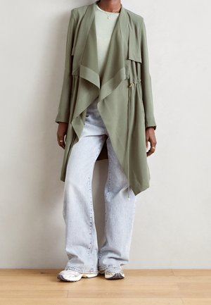 Personne portant une veste drapée vert olive sur un pull clair, un jean large bleu clair et des baskets beiges, debout sur un sol en bois.