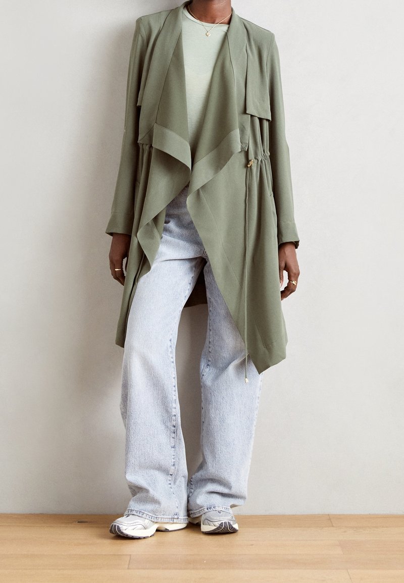 Personne portant une veste drapée vert olive sur un pull clair, un jean large bleu clair et des baskets beiges, debout sur un sol en bois.