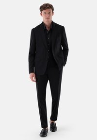 Schwarzer Anzug mit schmalem Schnitt, einreihiger Jacke und passenden Hosen. Getragen über einem schwarzen Hemd, kombiniert mit schwarzen Schuhen.