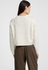 Maglione bianco lavorato a maglia con un motivo a treccia testurizzato, design accorciato e orlo a costine. Abbinato a pantaloni marroni, visto da dietro.