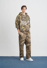 Conjunto de capuz e calças camufladas em tons de castanho e verde com um padrão de folhas. Apresenta um bolso frontal, mangas longas e cintura elástica. Sapatos brancos.