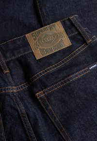 Donkerblauwe spijkerbroek met oranje stiksels, met een bruine "Superdry Standard Denim Goods" label boven de achterzak en een klein merkje.