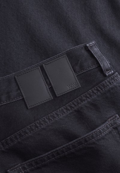 Mustmustekangas esitleb kahte ristkülikukujulist nahast sildiga vöökoht, millel on tikandiga aksessuaare ning tagatasku peente detailidega.