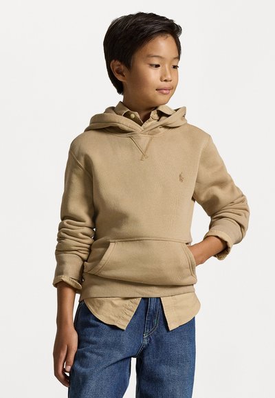 Sweatshirt bege com bolso canguru, punhos canelados e um logotipo no peito. Usado sobre uma camisa clara de colarinho e combinado com jeans azuis.