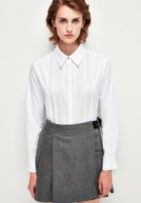 Camisa blanca de cuello con detalles de pliegues en el frente, acompañada de una falda gris de textura y cintura alta que presenta un cierre lateral.