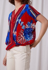 Blouse fleurie rouge avec des accents bleus, dotée d'un col en V, de manches courtes et d'une coupe ample. Détails avec des rayures et un motif texturé.