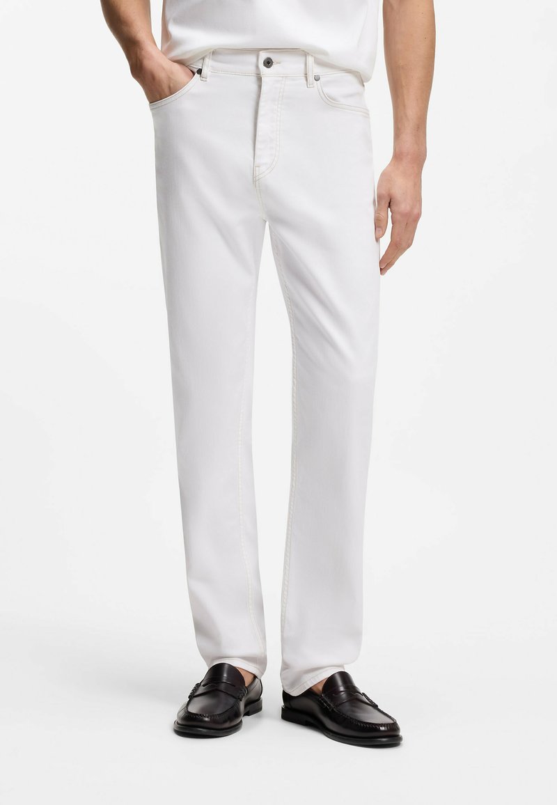 Personne portant un pantalon blanc droit avec poches avant, fermeture à boutons et glissière, associé à des mocassins en cuir noir.