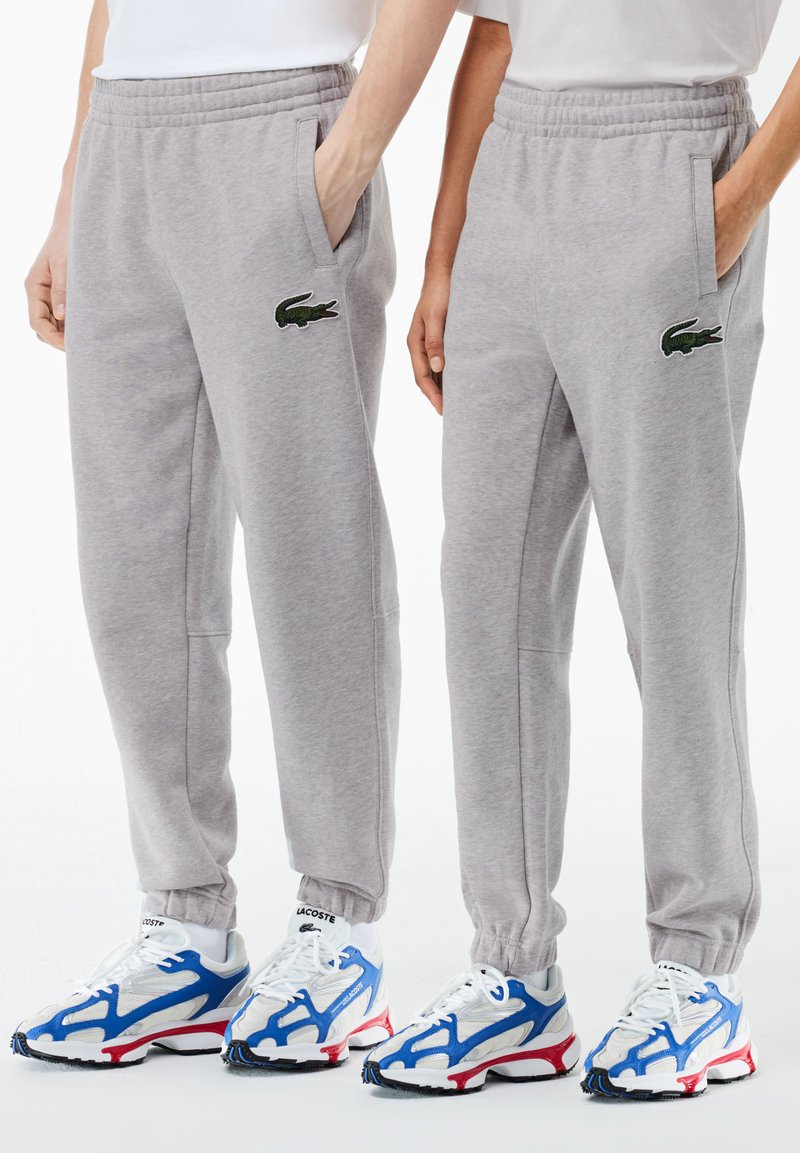 Dvoje ljudi nose svijetlosive trenirke s Lacoste krokodil logom, bijele Lacoste čarape i bijele tenisice s plavim i crvenim detaljima.